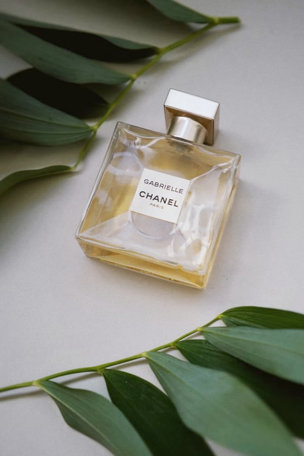 A picture of a bottle of perfume laying on a white surface.  There are leaves on two sides of the bottle. The bottle has a yellow liquid in it and is adorned with the words 'Chanel' and 'Gabrielle'.