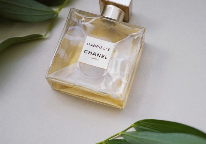 A picture of a bottle of perfume laying on a white surface.  There are leaves on two sides of the bottle. The bottle has a yellow liquid in it and is adorned with the words 'Chanel' and 'Gabrielle'.